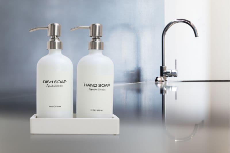 Frosted dish and hand soap pumps on a dark reflective counter near a faucet.
